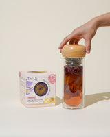 A hand placing the lid on the Paradise Double Wall Glass Bottle containing colorful flower tea.