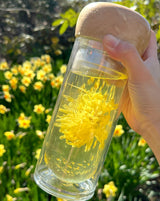 Person holding a Paradise Double Wall Glass Bottle with yellow flower tea in a garden setting.
