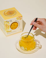 Hand stirring a cup of Royal Chrysanthemum tea beside the Iconic Bloom Trio set packaging.
