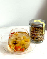 A clear glass teacup filled with Wild Chrysanthemum tea and a jar of Single Origin Wild Chrysanthemum on a marble table.