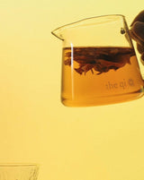 Small glass pot holding infused flower tea with a hand pouring against a warm yellow backdrop.