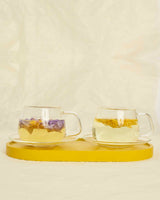 Glass cups & saucers (2) on a yellow tray with herbal tea and flower petals, perfect for tea rituals.