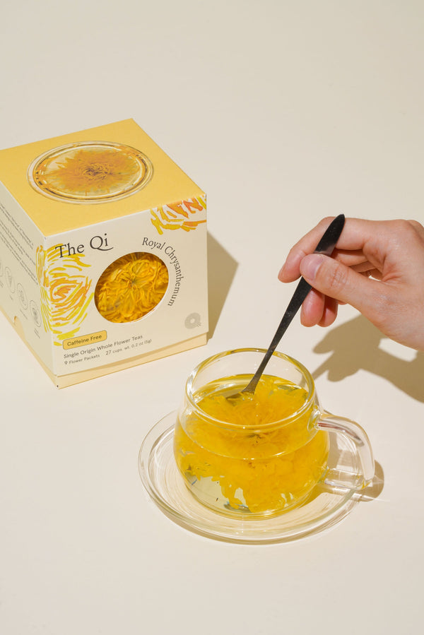 Hand stirring a cup of tea next to a box of The Qi Royal Chrysanthemum, representing wellness.